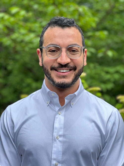 The image shows a man wearing glasses, smiling, with short hair, dressed in a button-up shirt and standing outdoors.