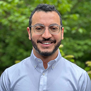A man with glasses and facial hair, smiling and wearing a button-up shirt, poses for a photo.