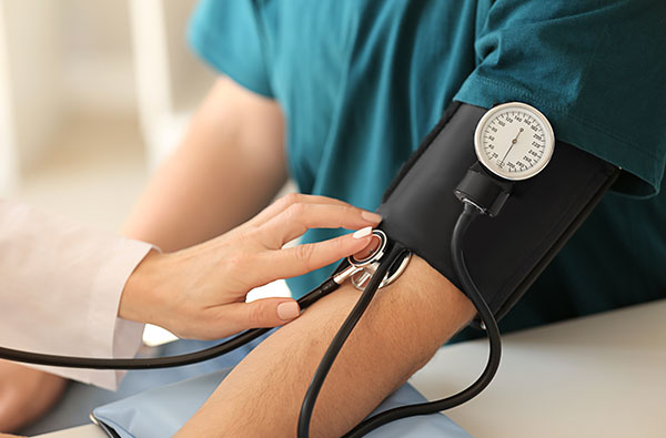 The image shows a healthcare professional taking a patient s blood pressure using a cuff while the patient sits on a chair, with the cuff wrapped around the patient s arm and the professional holding the device.