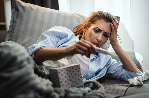 A woman laying on a couch with her hand on her head, looking distressed while holding an open box of tissues.