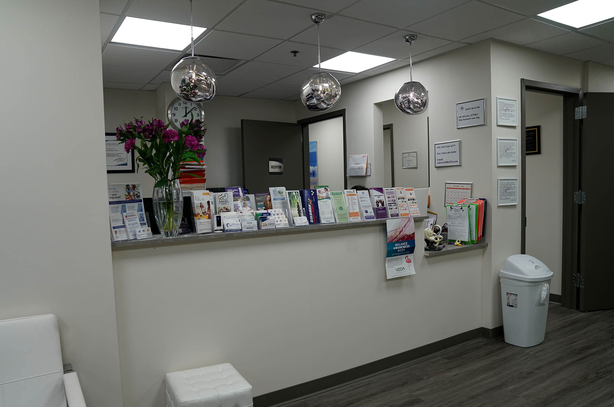 The image shows an interior view of a waiting room with a reception desk, a counter displaying various items, a shelf with brochures, a wall-mounted light fixture, and decorative elements such as vases with flowers.