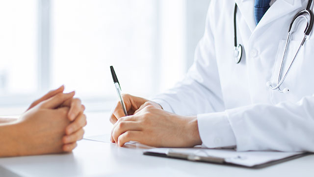 The image shows a medical professional sitting at a desk with a patient s hands resting on their lap, both wearing masks and looking towards each other, with the professional holding a pen and paper, suggesting a consultation or examination in progress.